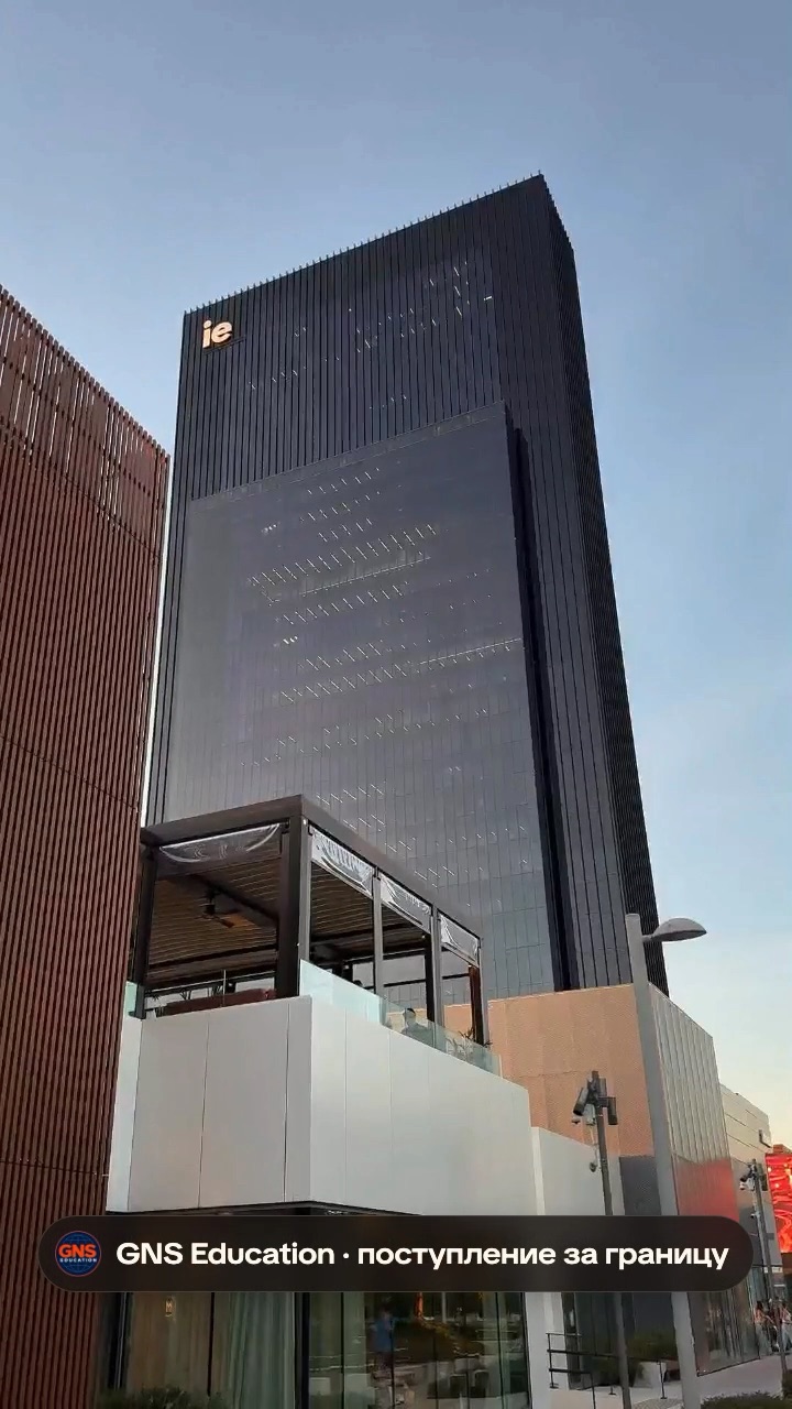 Тёмная башня IE Tower с логотипом ie на фасаде на фоне вечернего неба; у её подножия низкое белое здание с террасой под навесом