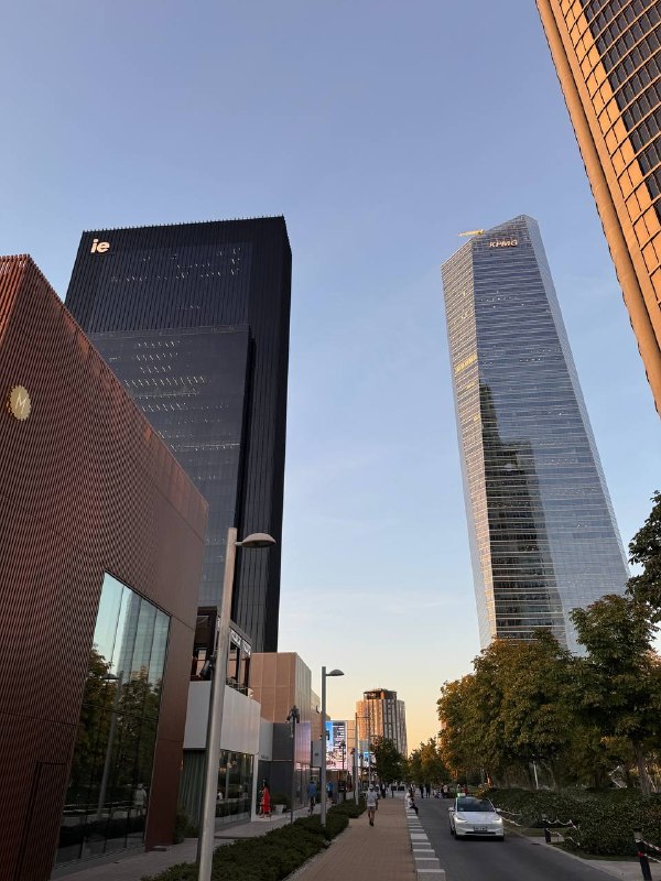 Мадрид на закате: слева тёмная башня IE Tower с логотипом ie, справа стеклянный небоскрёб с вывеской KPMG, между ними пешеходная аллея с деревьями